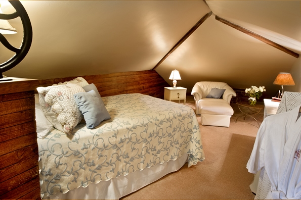 Large alcove area with full sized bed in foreground, with sitting chair, small table and table lamp in background.