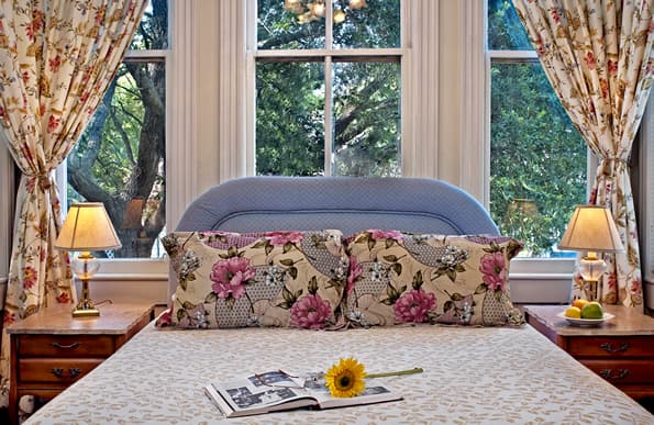 Bed with floral pillows.  Antique Nightstands and lamps on sides.  Floral Curtains on windows behind bed.