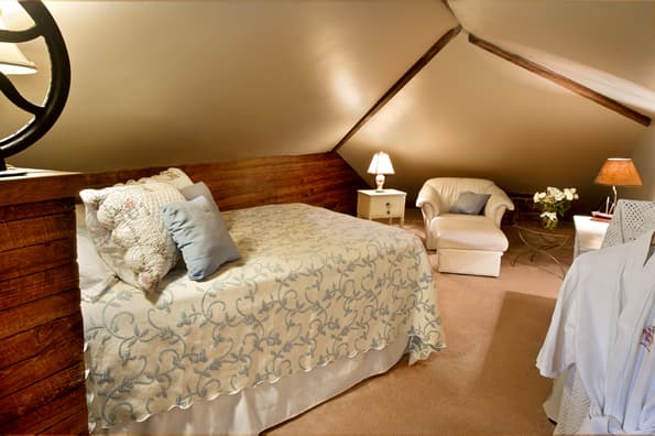 Large alcove area with full sized bed in foreground, with sitting chair, small table and table lamp in background.