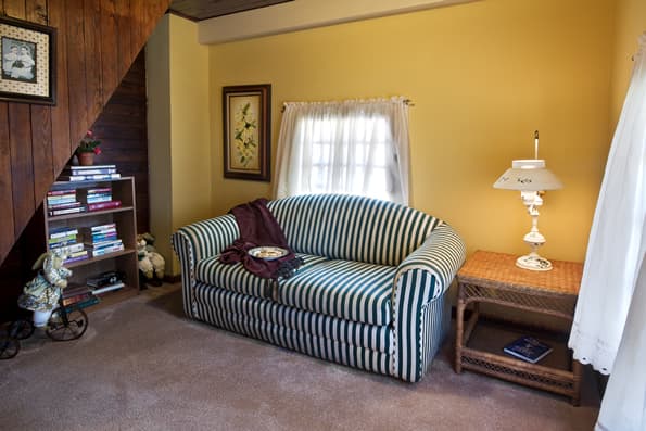 Nook with small loveseat, side table and table lamp for reading.  Bookcase and books to the side, under staircase.