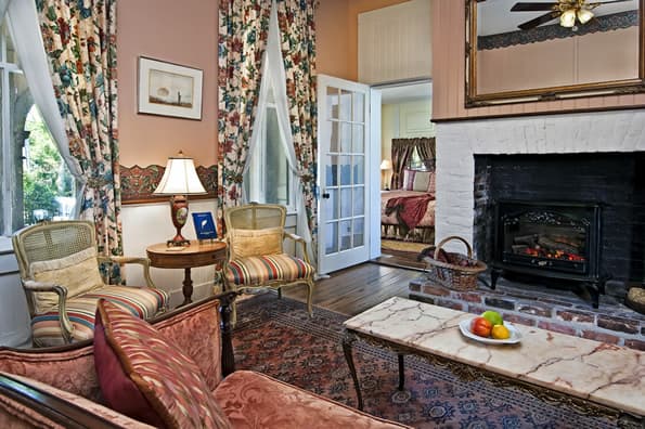 Living area with couch in foreground and large brick fireplace with stove insert in background.  Antique Table and lamp between windows and sitting chairs.