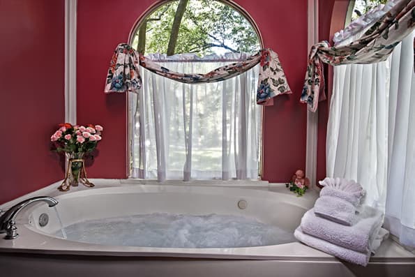 Large, jetted soaking tub with windows and sheer curtains.  Roses in vase on side of tub