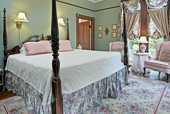King sized Colonial 4 poster bed with 2 highback chairs in background. Small table and lamp in between chairs, near windows.