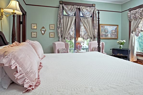 King sized colonial 4 poster bed with 2 highback chairs in background. Floral curtains on windows and paintings on walls.