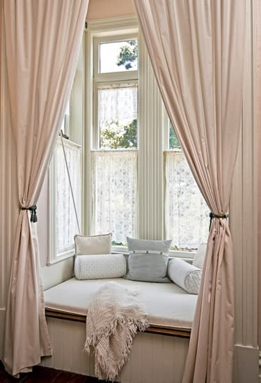 Bench seating area with pillows in window box. Sheer curtains on windows and Long pink blackout curtains held back by hooks
