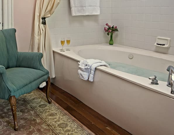 Large jetted soaking tub, with green highback chair to the side. 2 filled champagne flutes and a small vase with roses on the edge of the tub