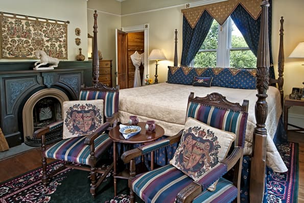 2 Highback chairs in front of King sized colonial 4 poster bed. Carved soapstone fireplace with tapestry is to the side. in the corner is an open closet door, revealing a cedar lined closet.