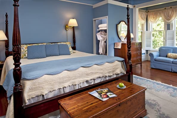 King sized Colonial 4 poster bed with antiques dresser in background. Cedar Chest at foot of bed. Blue Sofa in window box in background.