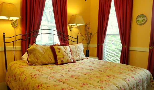 Floral pattern on king sized bed contrast with floral patterned window curtains.  Nightstand and table lamps on the side of the bed.