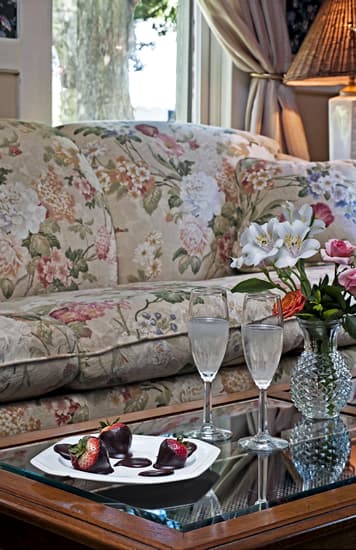 Closeup view of coffee table with flowers in vase, and 2 filled champagne flutes and chocolate dipped strawberries in front of Floral sofa.