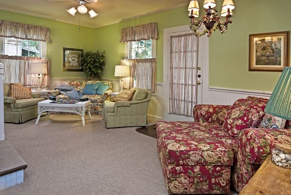 Living room with oversized red chair in corner. seating area in background features 2 chairs, a wicker coffee table and a small Floral sofa in background. Walls are green and there are 2 hanging lights and a ceiling fan.