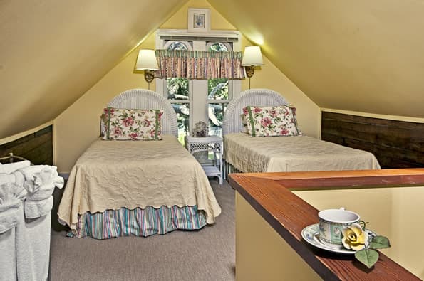 two twin beds with wicker headboard are separated by a wicker nightstand. Mounted lamps above headboards. Roofline angles down towards side walls.