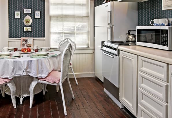 Wicker Kitchen table in kitchen has bowl of fruit and 4 place settings. Cabinets, microwave, Stove and Refridgerator are on side wall, with window in background, behind table.