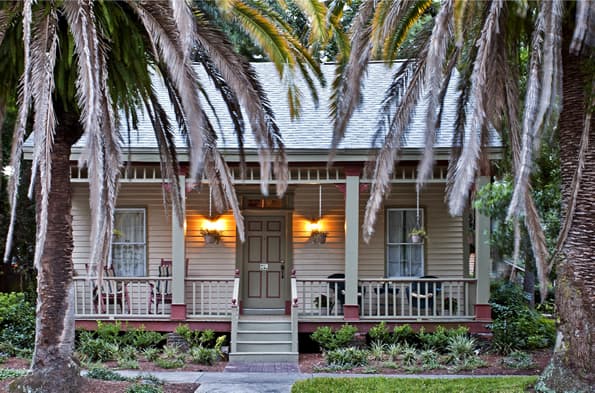 Front porch shows Chairs and side tables on either side on overhanging porch entry. Palm trees are framing either side.