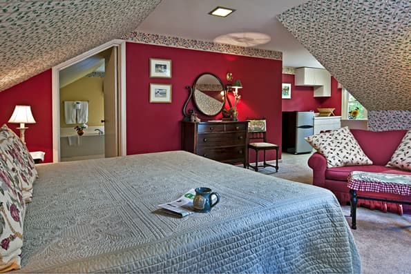 King sized bed with coffee cup and newspaper on bed in foreground. Red walls, antique dresser and matching red sofa in background. Kitchen sink and small refridgerator visible further back.