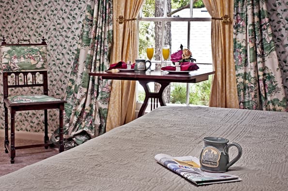 Coffee cup and newspaper on bed in foreground. Small table by window with 2nd coffee cup, and 2 mimosas in Champagne flutes in background.