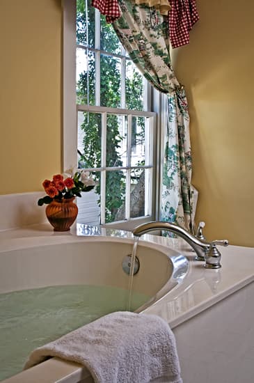Large jetted soaking tub with a small vase with roses on the edge of the tub and window with flowral curtains in background.
