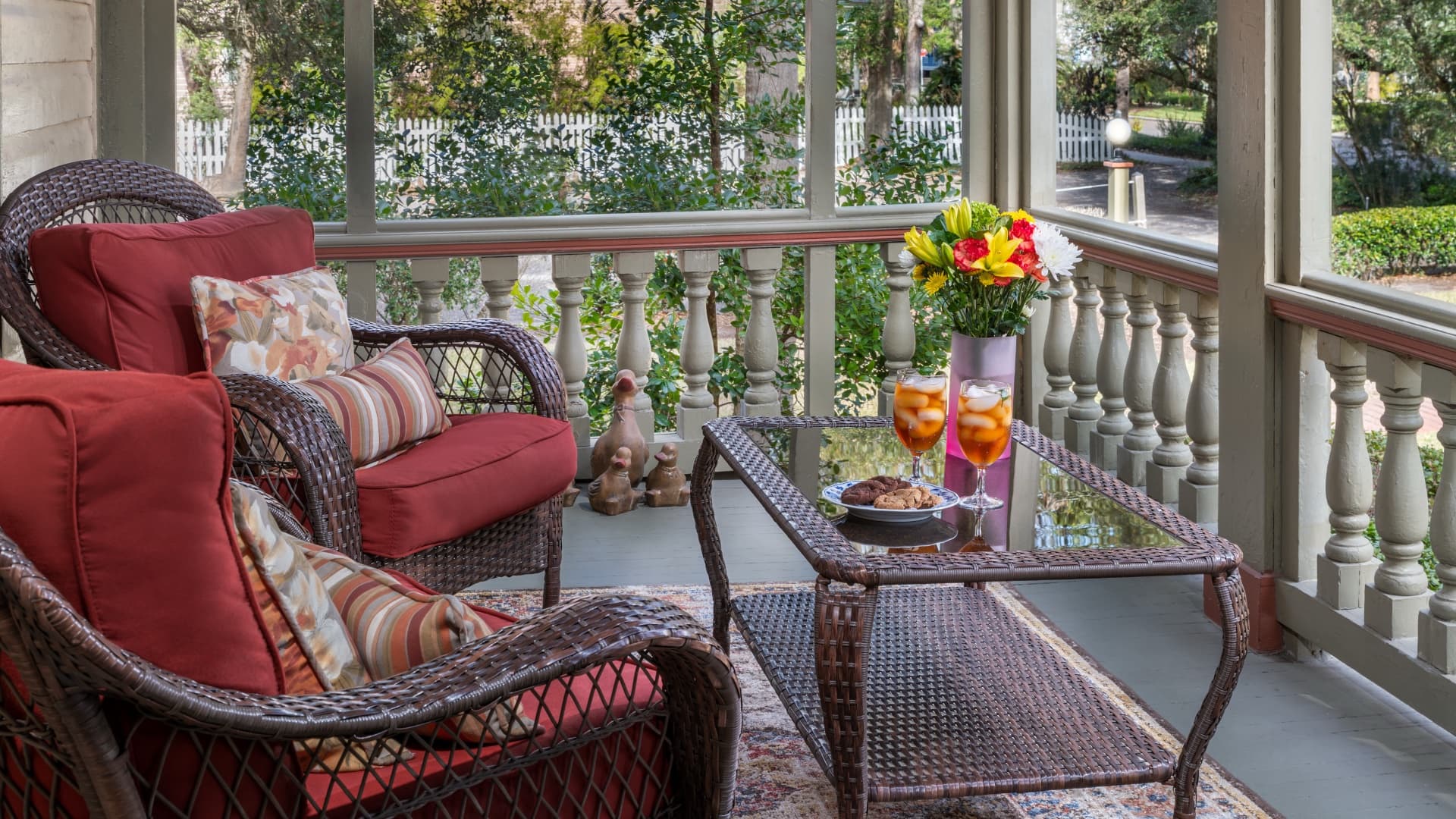 A cozy porch features wicker furniture, vibrant pillows, a glass table with drinks and cookies, and a flower vase, surrounded by greenery.