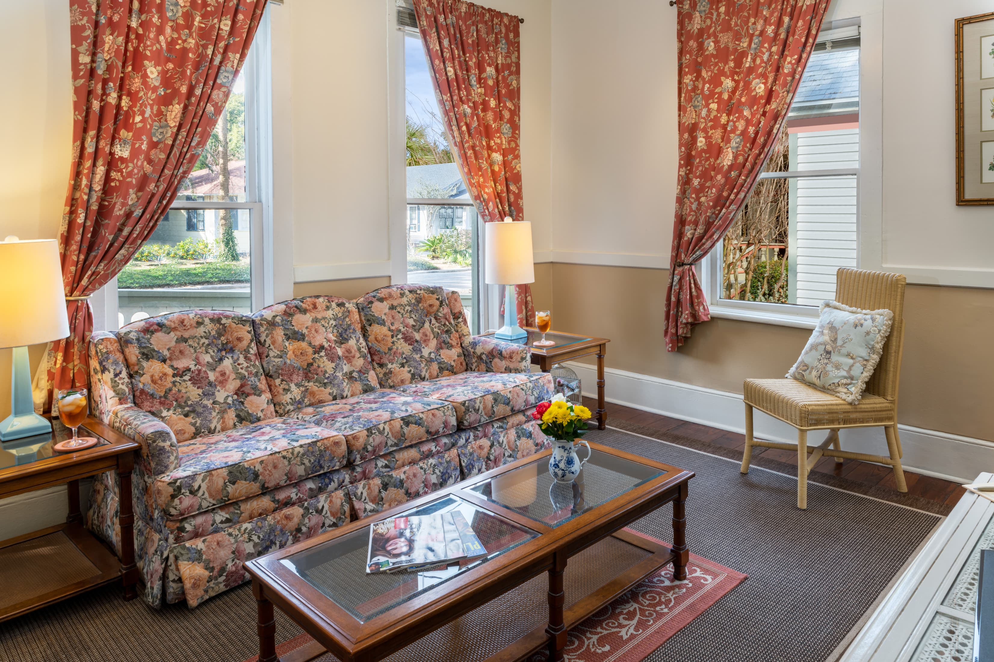 Cozy cottage-style living room with floral sofa, vintage furnishings, hardwood floors, and large windows with decorative curtains.
