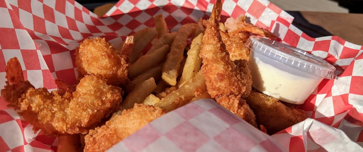 A serving of crispy fried shrimp and French fries with a side of dipping sauce in a checkered paper basket.