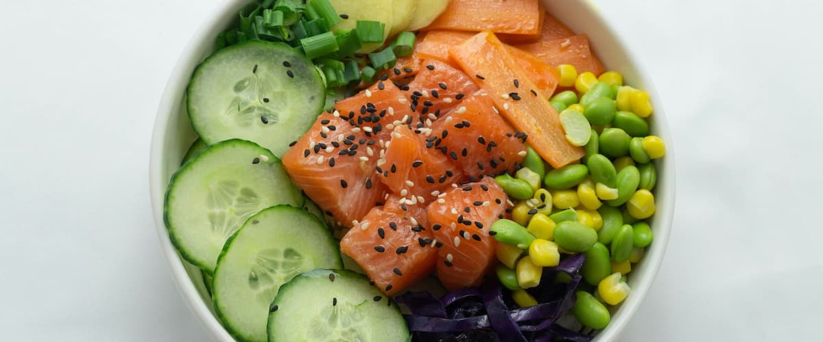 A colorful bowl featuring diced salmon, cucumber slices, edamame, corn, and purple cabbage.