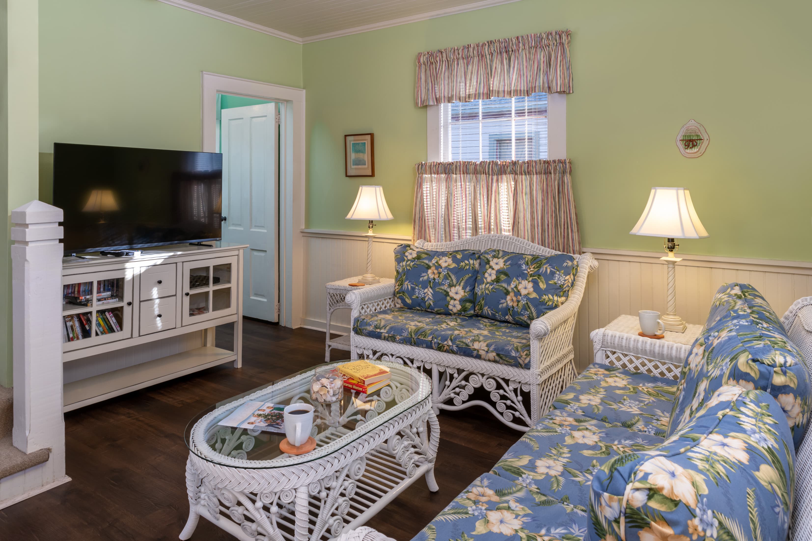 Bright coastal-style living room featuring wicker seating with floral cushions, hardwood floors, large flat-screen TV, and relaxing cottage décor.