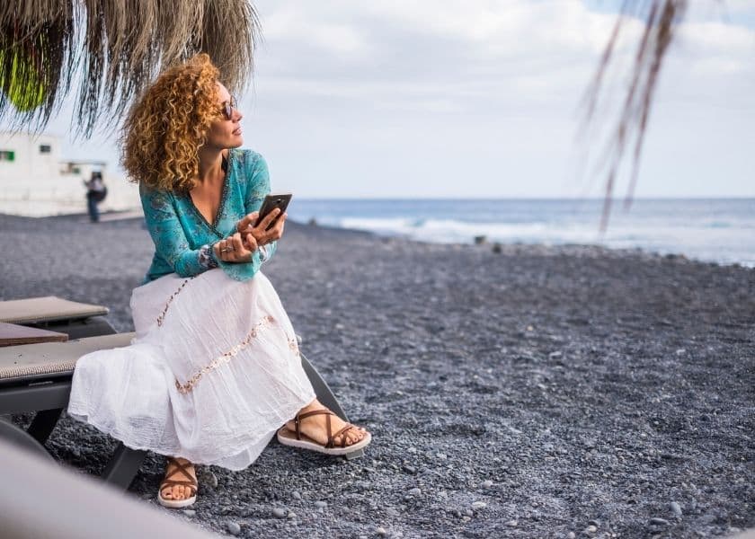 A woman in a turquoise top and white skirt sits on a beach chair, looking at her phone with the ocean in the background. A woman in a turquoise top and white skirt sits on a beach chair, looking at her phone with the ocean in the background.