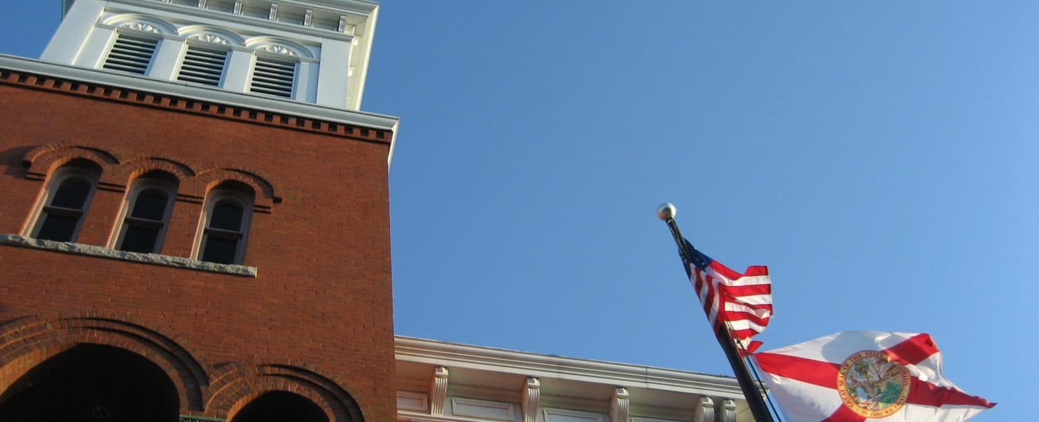 A red brick building with a clock tower displays the American and Florida state flags against a clear blue sky.