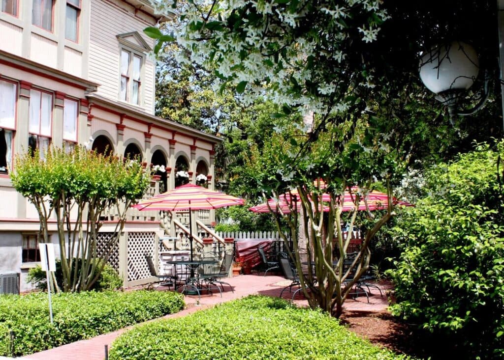 Victorian-style house with red and white striped umbrellas over patio tables surrounded by lush greenery.