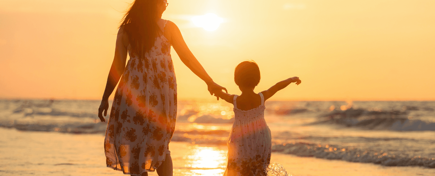 A woman and a girl walk hand-in-hand along the beach at sunset.