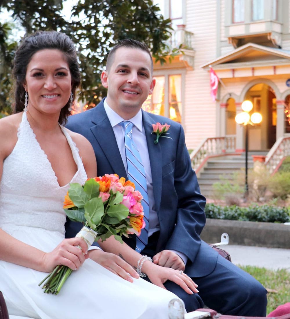 A smiling bride and groom sit together, holding a colorful bouquet, with a historic house in the background.