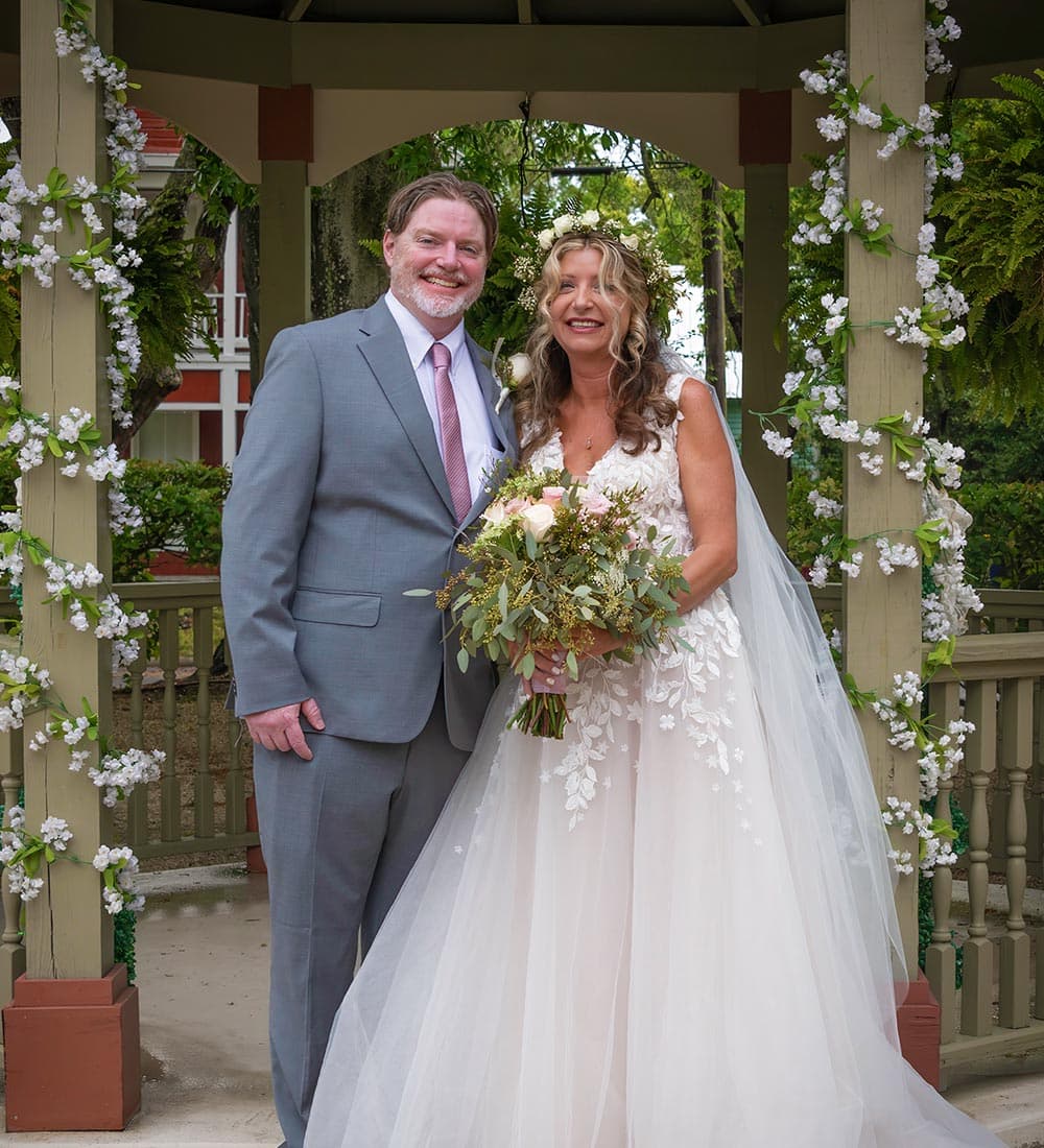 A smiling couple stands together under a floral-adorned gazebo, with the bride holding a bouquet and wearing a flowing wedding dress.