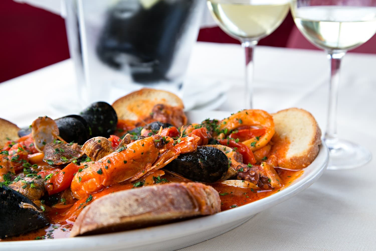A plate of seafood pasta in a rich tomato sauce, garnished with parsley, accompanied by toasted bread and two glasses of white wine.