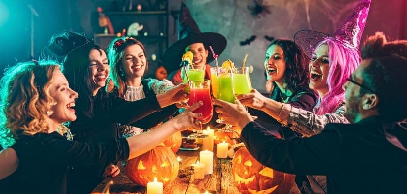 A group of eight friends in Halloween costumes joyfully toast with colorful drinks surrounded by carved pumpkins and candles.