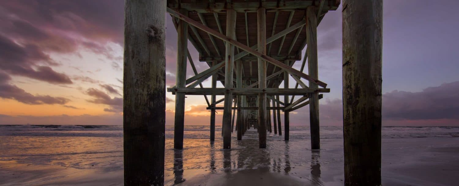 View from beneath a wooden pier at sunset, reflecting on calm water.