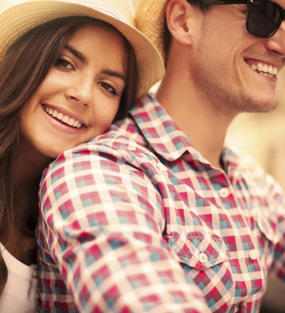 A smiling woman leans affectionately on a man wearing sunglasses, both enjoying a sunny day.