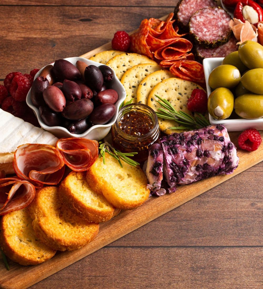 A wooden platter featuring an assortment of meats, cheeses, olives, crackers, and garnishes.