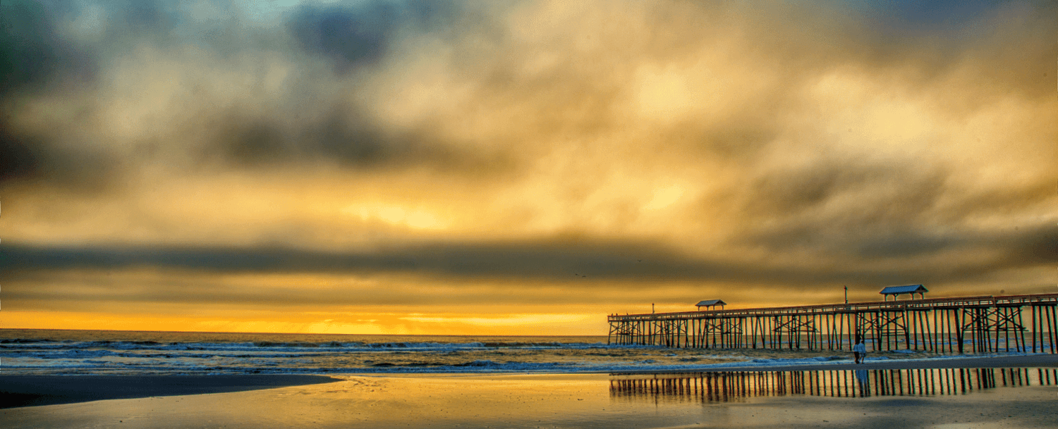 A wooden pier stretches over a reflective beach under a dramatic orange and gray sky at sunset.