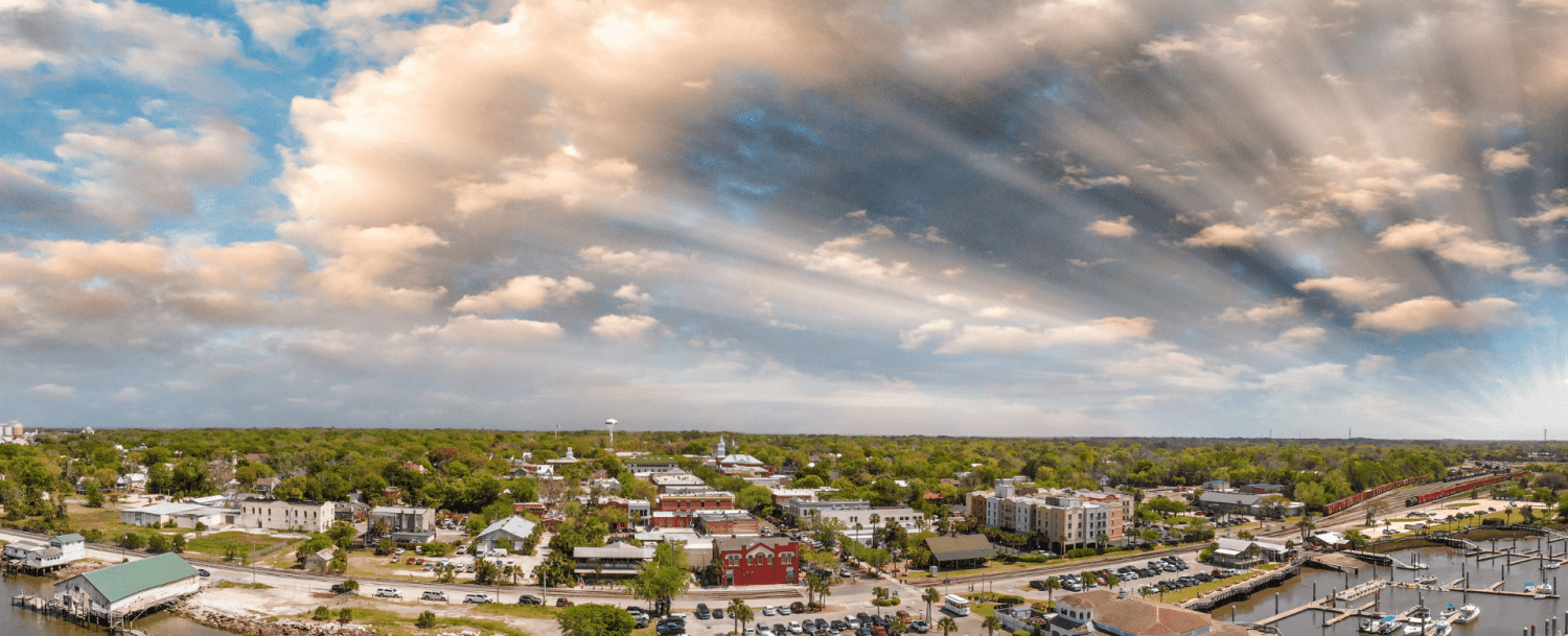 A panoramic view of a coastal town with lush greenery, colorful buildings, and a marina under a dramatic sky.