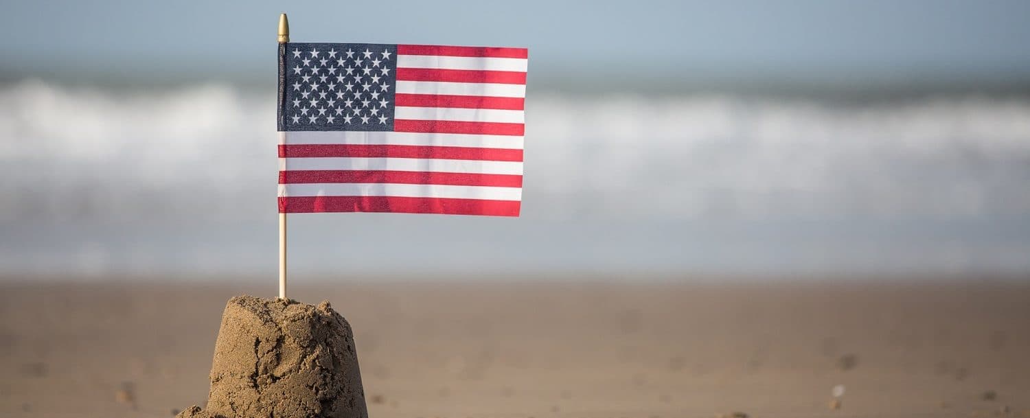 A small American flag is planted in a sandcastle on a beach.