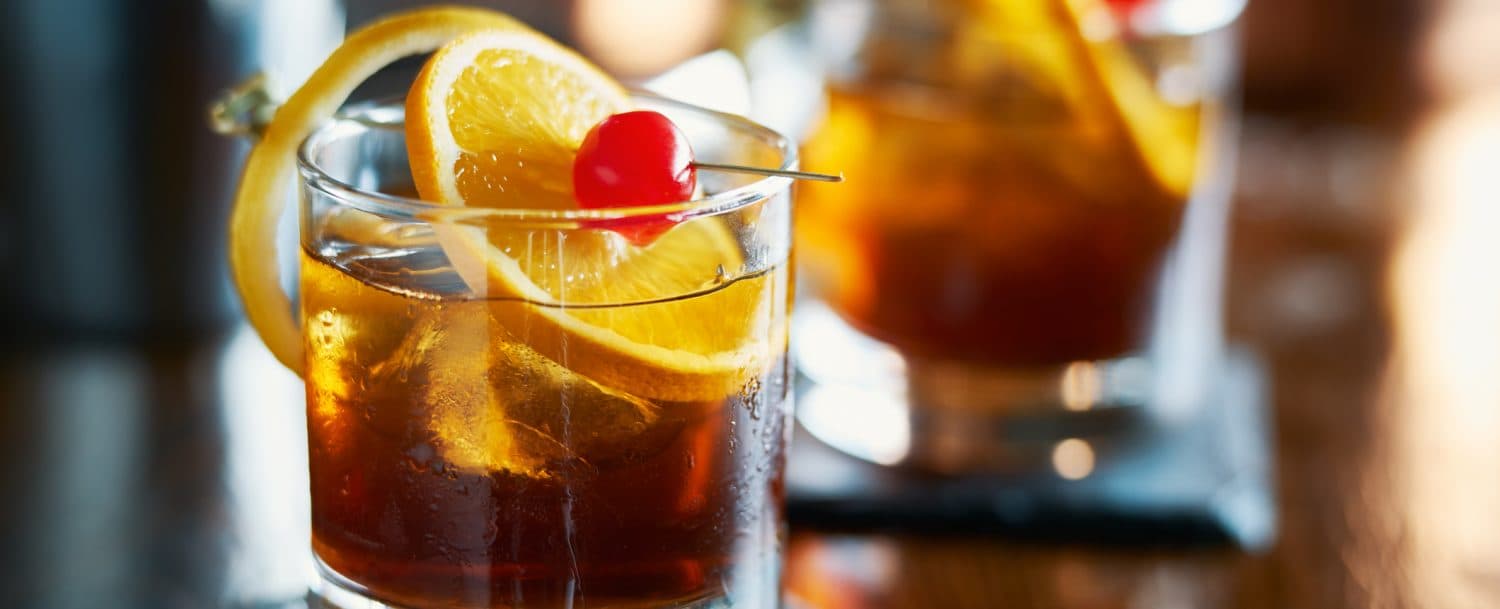 A close-up of a cocktail glass filled with a dark drink, garnished with lemon slices and a cherry.