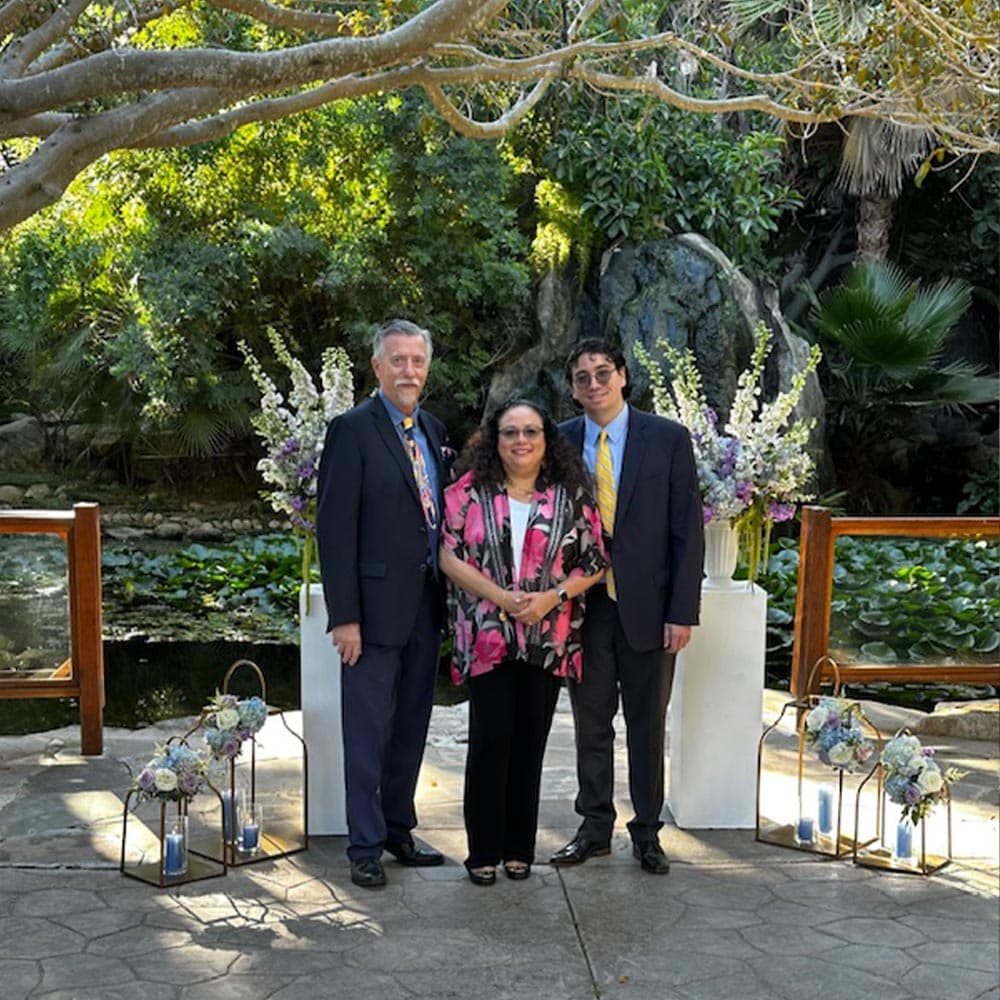Three individuals pose for a photo in a lush garden setting with floral arrangements and water features in the background.