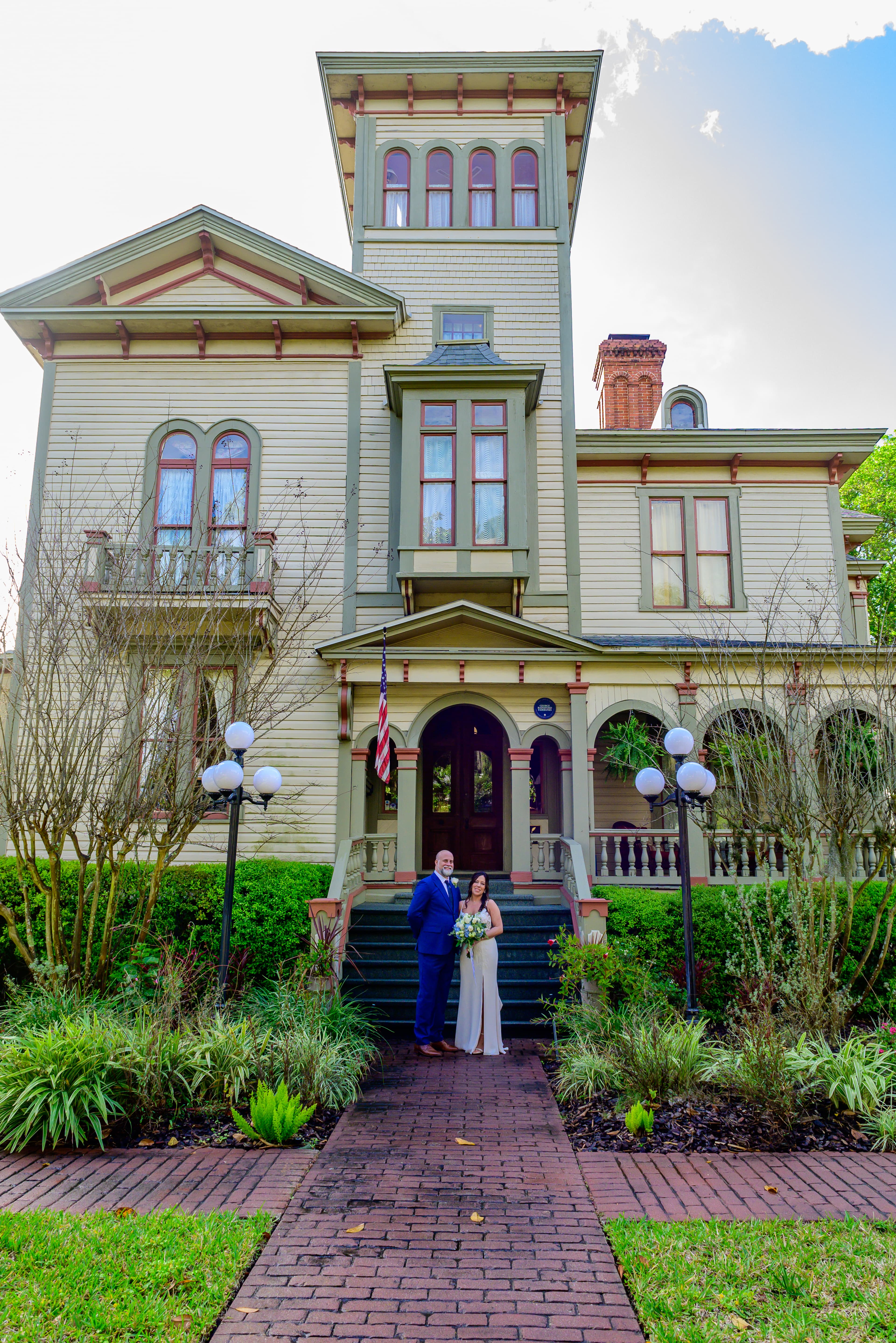 A couple stands in front of a large Victorian-style house, smiling and holding flowers.