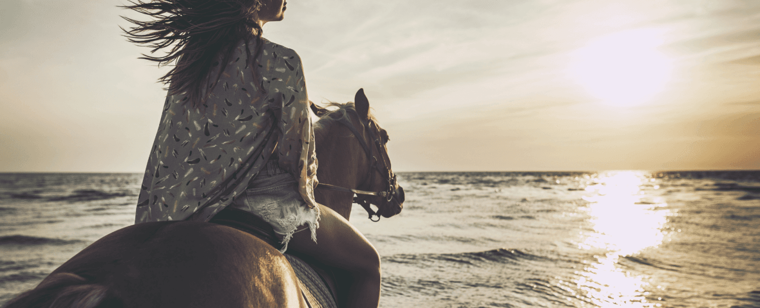 A woman rides a horse along the beach at sunset.