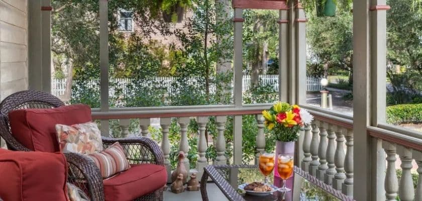 A cozy porch with red cushions, a floral centerpiece, and drinks set on a table surrounded by greenery.