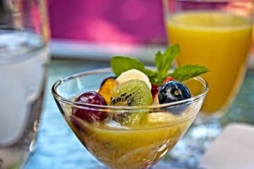 A glass bowl filled with assorted fresh fruit, accompanied by a glass of orange juice and water in the background.