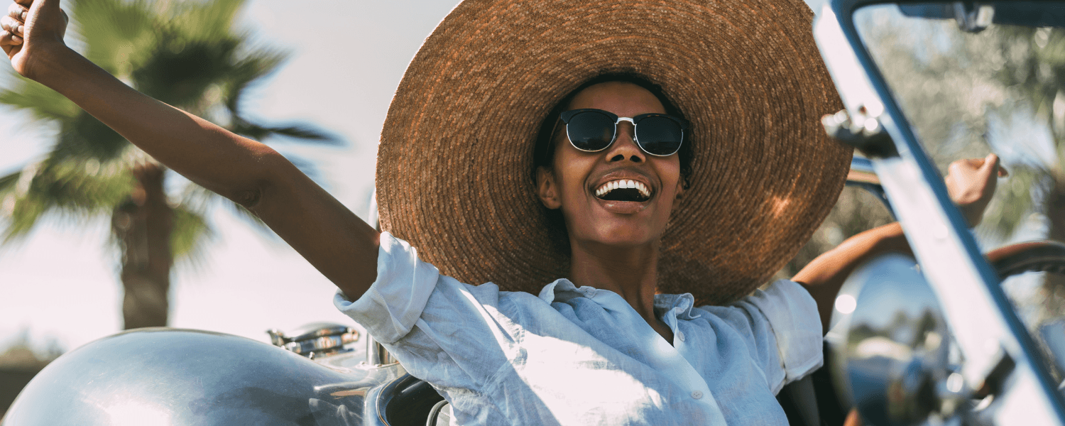 A woman in a large straw hat and sunglasses joyfully leans out of a vintage car against a sunny backdrop.