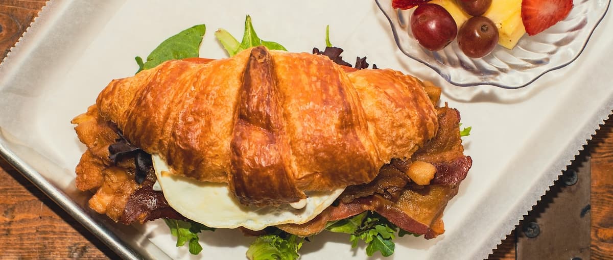 A croissant sandwich filled with bacon, fresh greens, and a fried egg, served with a side of assorted fruit.
