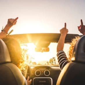 Two people in a convertible raise their arms joyfully against a sunset backdrop.