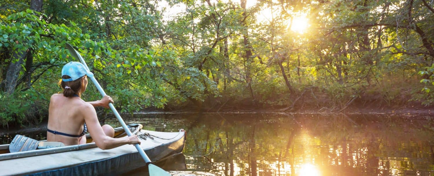 A person paddles a kayak on a serene river surrounded by trees at sunset.